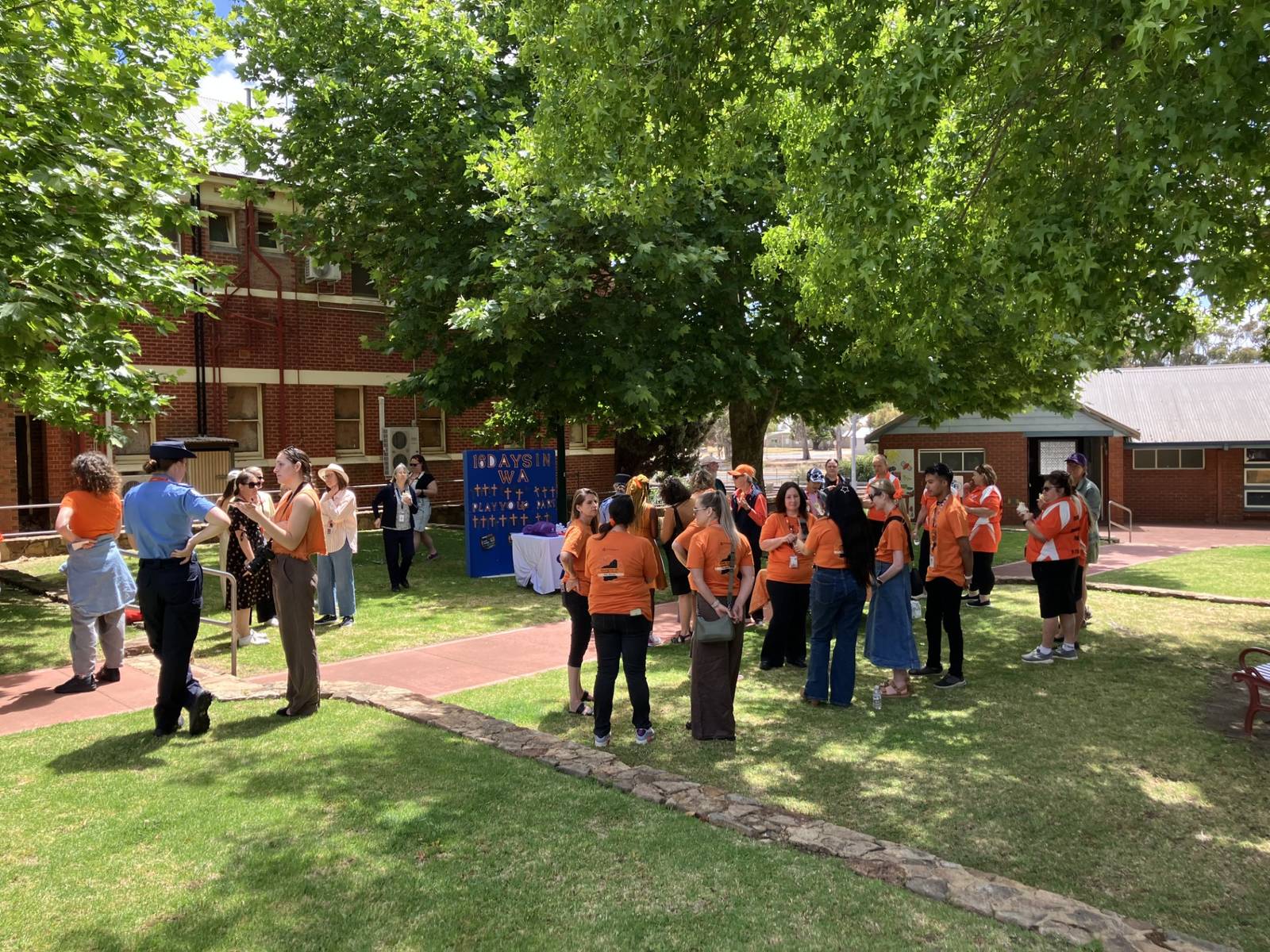 Community members wearing orange gather outside a historic red-brick building in Narrogin for the 16 Days in WA march, raising awareness about family and domestic violence.